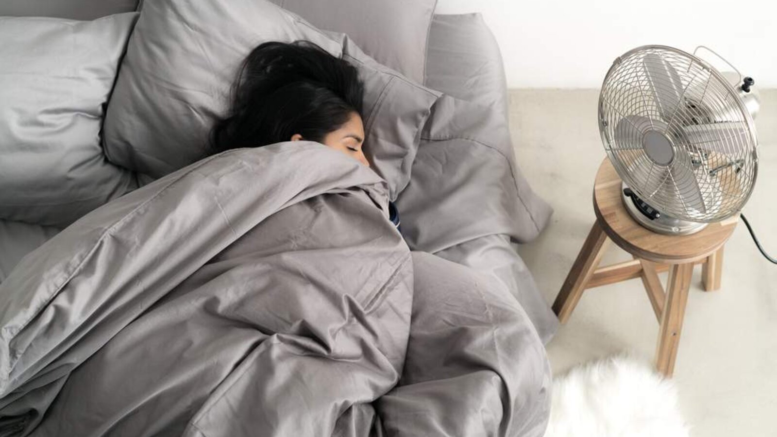A person sleeping comfortably in cool gray sheets with a fan running nearby